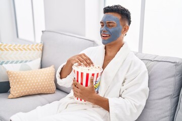 Wall Mural - Young latin man relaxed on sofa with facial mask treatment eating popcorn at home