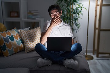 Sticker - Young hispanic man using laptop at home at night laughing and embarrassed giggle covering mouth with hands, gossip and scandal concept