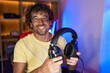 © Krakenimages.com - Young hispanic man streamer smiling confident holding headphones at gaming room