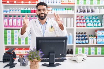 Sticker - Hispanic man with beard working at pharmacy drugstore looking at the camera smiling with open arms for hug. cheerful expression embracing happiness.