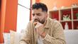 Young hispanic man eating doughnut sitting on sofa at h