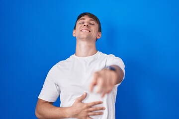 Sticker - Caucasian blond man standing over blue background laughing at you, pointing finger to the camera with hand over body, shame expression