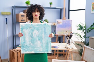 Poster - Young brunette woman with curly hair holding painter canvas relaxed with serious expression on face. simple and natural looking at the camera.