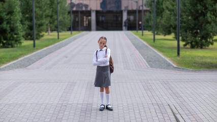 Wall Mural - A sad caucasian girl in uniform and with a backpack holds her arms crossed on her chest stands outdoors after school.