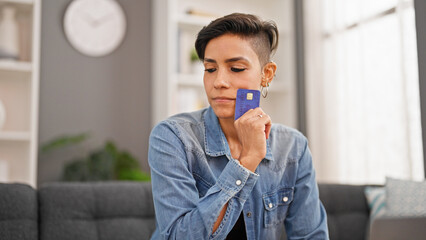 Young beautiful hispanic woman holding credit card sitting on sofa thinking at home