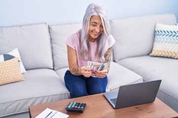 Wall Mural - Middle age grey-haired woman using laptop counting dollars at home