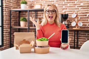 Canvas Print - Blonde woman eating take away food showing smartphone screen celebrating victory with happy smile and winner expression with raised hands