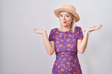 Poster - Young caucasian woman wearing flowers dress and summer hat clueless and confused expression with arms and hands raised. doubt concept.