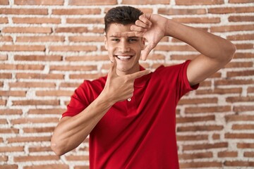 Sticker - Young hispanic man standing over bricks wall smiling making frame with hands and fingers with happy face. creativity and photography concept.