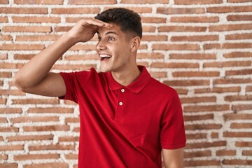 Sticker - Young hispanic man standing over bricks wall very happy and smiling looking far away with hand over head. searching concept.