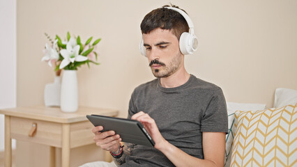 Wall Mural - Young hispanic man using touchpad and headphones sitting on bed at bedroom