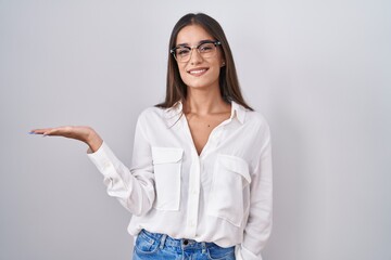 Canvas Print - Young brunette woman wearing glasses smiling cheerful presenting and pointing with palm of hand looking at the camera.