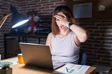 Sticker - Brunette woman working at the office at night peeking in shock covering face and eyes with hand, looking through fingers with embarrassed expression.