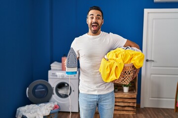 Poster - Young hispanic man with beard holding iron and clothes at home celebrating crazy and amazed for success with open eyes screaming excited.