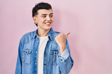 Poster - Non binary person standing over pink background smiling with happy face looking and pointing to the side with thumb up.