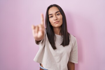 Wall Mural - Young hispanic woman standing over pink background pointing with finger up and angry expression, showing no gesture
