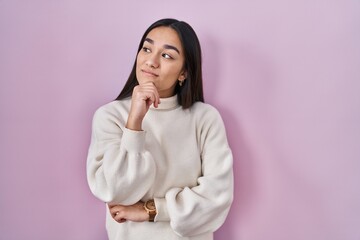 Wall Mural - Young south asian woman standing over pink background with hand on chin thinking about question, pensive expression. smiling with thoughtful face. doubt concept.