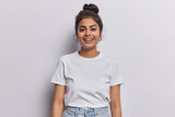 Fototapeta  - Horizontal shot of Indian woman with dark hair gathered in bun smiles pleasantly being in good mood dressed in casual t shirt and jeans isolated over white background. People and emotions concept