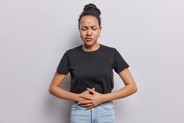 Unhappy dark haired woma keeps hands on belly suffers from stomach ache discomfort or menstrual cramps has eyes closed dressed in casual clothes isolated over white background. Health problems