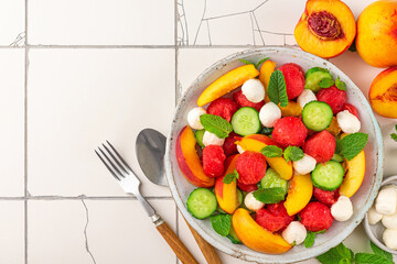 Wall Mural - Summer fruit salad with mozzarella cheese, cucumber and mint in a plate with cutlery on white tile table. top view
