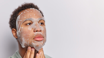 Wall Mural - Horizontal shot of adult woman applies sheet beauty mask on face showcases his dedication to skin care keeps hands under chin concentrated aside isolated over white background copy space on right side