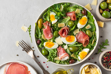 Wall Mural - Prosciutto salad with parmesan, olives, eggs and arugula in a plate on gray background. Italian food. top view