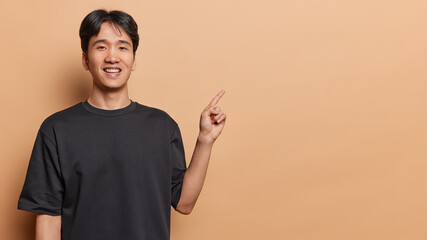 Positive dark haired Japanese man pointing away towards copy space for your text or logo smiles toothily dressed in casual black t shirt isolated on brown background. People and advertisement concept