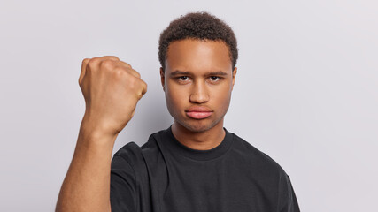 Wall Mural - Angry dark skinned adult man clenches fist expresses negative emotions demonstrates his irritation dressed in casual black t shirt isolated over white background. People and annoyance concept