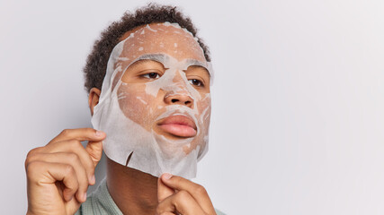 Wall Mural - Image of serious black African guy concentrated aside attentively applies nourishing sheet mask on face takes care of skin isolated over white background copy space for your promotional content