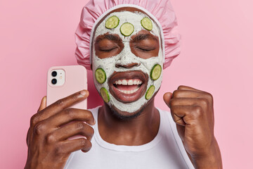 Overjoyed dark skinned man clenches fist celebrates good news holds mobile phone poses with beauty mask and fresh cucumber slices in face wears bath hat white t shirt isolated over pink background