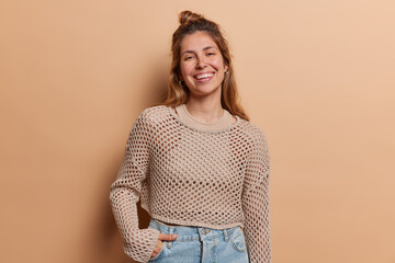 Poster - Positive young European woman stands carefree and happy against brown background keeps hand in pocket of jeans wears cropped knitted jumper smiles broadly isolated. People and lifestyle concept