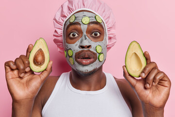 Stunned young man applies beauty mask with cucumbers to face holds halves on avocado uses natural cosmetic products wears bath hat and white t shirt isolated over pink background. Wellness concept