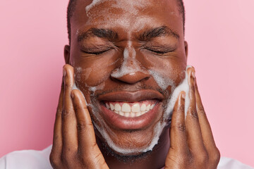 Close up shot of young African man exudes pure delight as he cleanses his face with soap pleased with condition of skin shows white teeth keeps eyes closed isolated over pink background. Daily hygiene