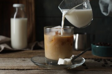 Pouring milk into glass with ice cubes and coffee at wooden table, closeup