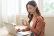 © KMPZZZ - Wellness and dieting asian young woman, girl working from home using computer, typing or searching prescription on medicine label about vitamins information online, holding bottle of food supplement.