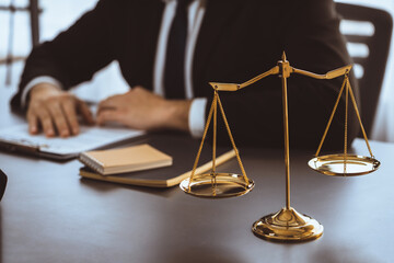 Wall Mural - Focus shiny golden balanced scale on blurred background of lawyer working in his desk at law firm office. Scale balance for righteous and equality judgment by lawmaker and attorney. Equilibrium