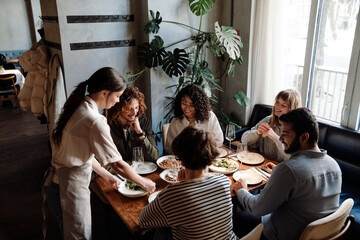 Wall Mural - Young waitress serving group of cheerful friends in restaurant