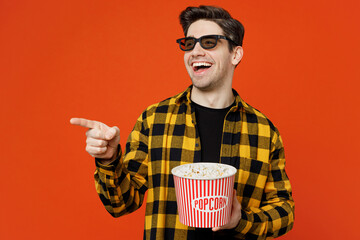 Poster - Young man in 3d glasses wears yellow checkered shirt black t-shirt watch movie film hold bucket of popcorn in cinema point index finger aside isolated on plain red orange background studio portrait.