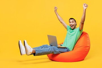 Wall Mural - Full body young IT man of African American ethnicity he wears casual clothes green t-shirt hat sit in bag chair hold use work on laptop pc computer raise up hands isolated on plain yellow background.