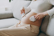 © ViDi Studio - Young pregnant woman wears casual clothes put little white socks on belly sits on grey sofa couch stay home hotel flat rest relax spend free spare time in living room indoor. Maternity family concept.