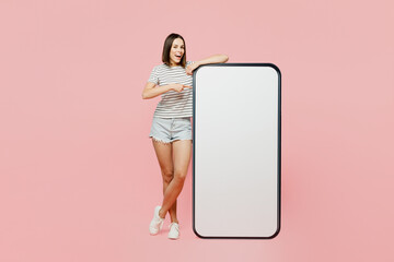 Full body young fun caucasian woman she wears casual clothes t-shirt point index finger on big huge blank screen mobile cell phone smartphone with area isolated on plain pastel light pink background.