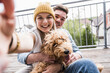 © Westend61 - Happy woman taking selfie with man and dog on balcony