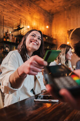 Vertical portrait of happy young woman paying bill with a contactless credit card in a restaurant. Cheerful female smiling holding a creditcard and giving a payment transaction to the cashier in a bar