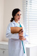 © DJ Creative Studio - Portrait beautiful Latin female doctor stands confidently by the window, preparing for patient consultations and working in the hospital.