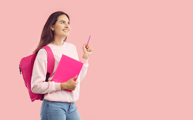 Smiling brunette student girl wearing pink sweatshirt and jeans with pink backpack and copybook looking up and pointing pencil up on copy space on pink background. Sincere emotions, advertisement.