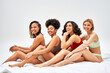 © LIGHTFIELD STUDIOS - Happy and multiethnic women in colorful lingerie smiling at camera while sitting together on white bed isolated on grey, different body types and self-acceptance concept, multicultural models