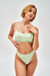 © LIGHTFIELD STUDIOS - Sexy young blonde woman with natural makeup in light green lingerie touching chest and looking away while standing isolated on grey, self-acceptance and body positive concept