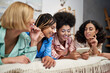 © LIGHTFIELD STUDIOS - Positive african american woman holding smartphone near blurred multiethnic girlfriends in colorful pajama lying on bed during pajama party in bedroom at home, bonding time in comfortable sleepwear