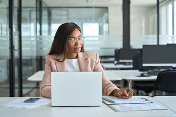 Busy professional young African American business woman company manager, female employee executive wearing suit working in office with laptop computer writing notes sitting at desk.