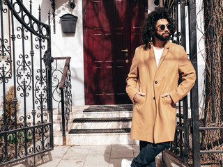 Handsome hipster model.  Unshaven Arabian man dressed in brown coat clothes. Fashion male with long curly hairstyle posing outdoors in street. Cheerful and happy. At sunny day